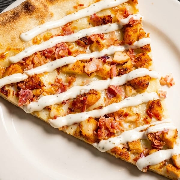 A slice of pizza with chicken, bacon, and a ranch drizzle on a white plate.