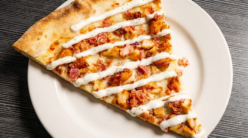 A slice of pizza with chicken, bacon, and a ranch drizzle on a white plate.