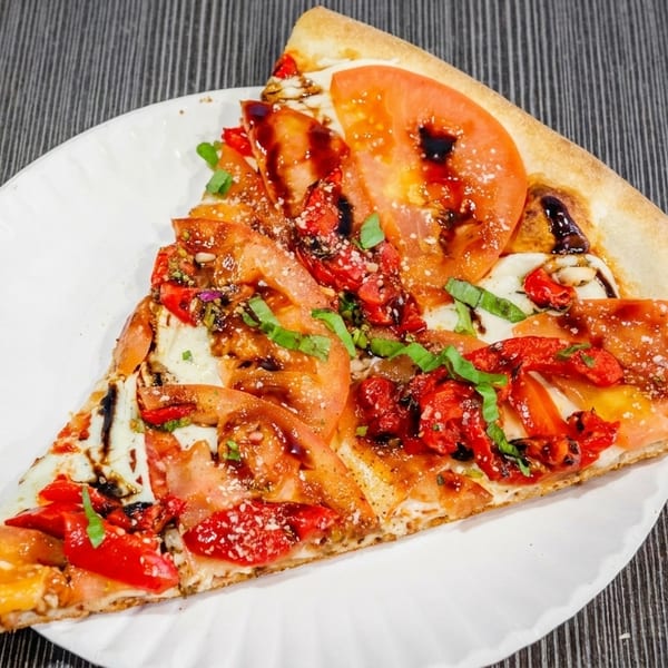 A large slice of pizza with tomatoes, red peppers, basil, and balsamic glaze on a white plate.