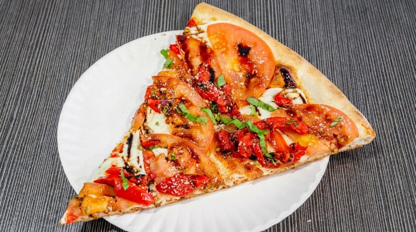 A large slice of pizza with tomatoes, red peppers, basil, and balsamic glaze on a white plate.