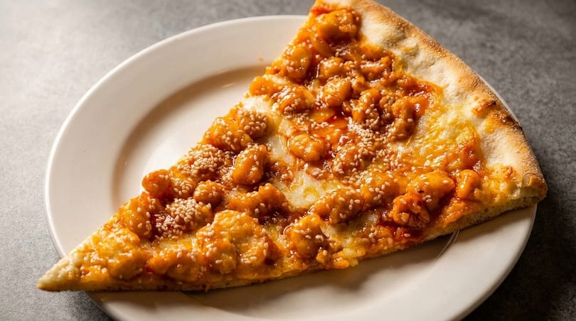 A slice of General Tso's chicken pizza, topped with sesame seeds, on a white plate.