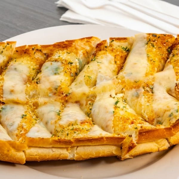 Cheesy garlic bread, cut into slices, on a white plate with napkins and cutlery in the background.