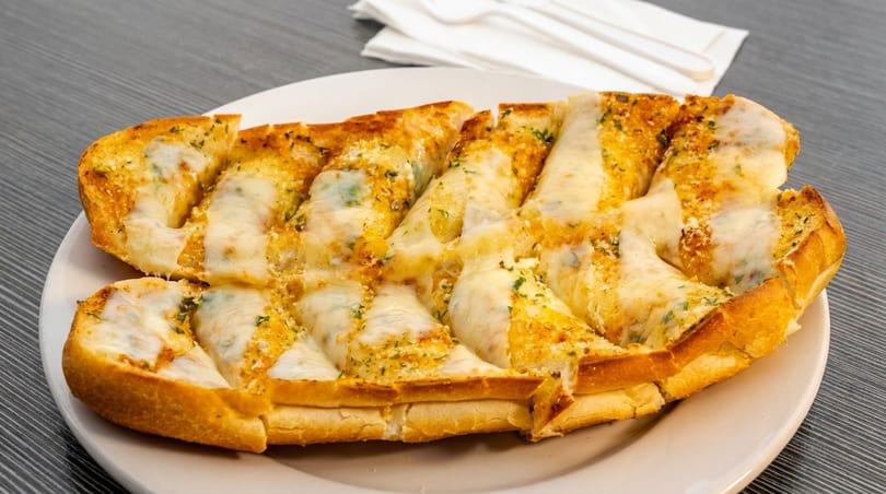 Cheesy garlic bread, cut into slices, on a white plate with napkins and cutlery in the background.