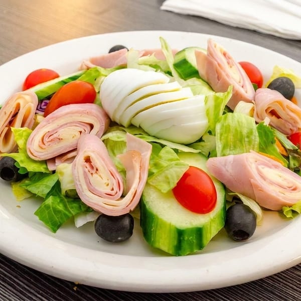 Chef salad with ham and cheese rolls, sliced egg, lettuce, tomatoes, cucumbers, and olives.