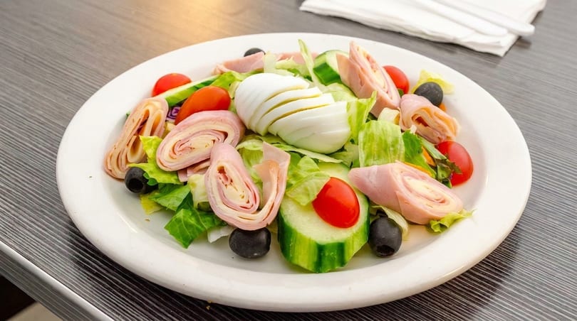 Chef salad with ham and cheese rolls, sliced egg, lettuce, tomatoes, cucumbers, and olives.
